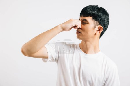 Photo for Unhappy young man, with a shocked expression, holds his nose due to a smelly and bad odor. Studio shot isolated on white, illustrating his strong reaction to the smell. - Royalty Free Image