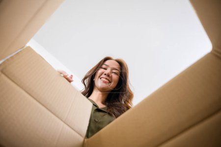 Téléchargez les photos : Happy Asian young woman opening carton box from internet store order shopping online at home, Smiling female surprised unpacking christmas gift big box and looking inside, inside bottom view - en image libre de droit
