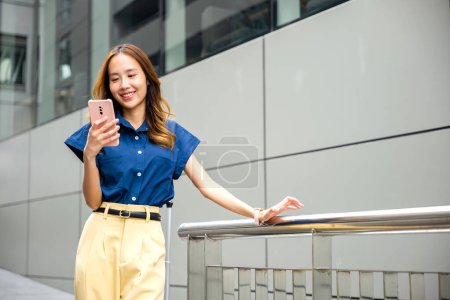 Photo for A young woman wearing blue shirt texting on her phone while strolling in the city. Girl in a good mood, busy with her smartphone app. - Royalty Free Image
