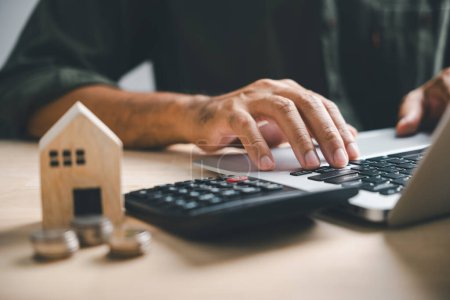 Photo for Hand presses calculators, pondering home refinance. Wooden house model, buy or rent note on desk. Saving for property purchase, mortgage payment strategy. Tax, credit analysis for financial success. - Royalty Free Image