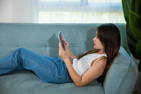 Téléchargez les photos : Happy Asian woman playing smart tablet at home while relaxing on sofa living room, Beautiful female sitting on couch using digital tablet pc, technology social media - en image libre de droit
