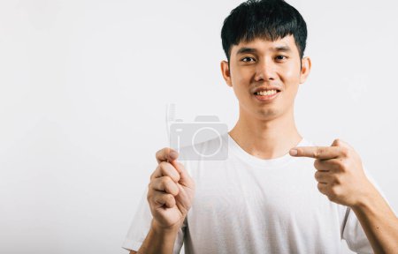 Foto de Un joven asiático sonríe mientras sostiene un cepillo de dientes y se lava los dientes, señalando el cepillo. Estudio aislado sobre fondo blanco, presentando salud dental con expresión positiva. - Imagen libre de derechos