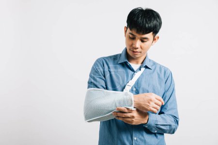 Photo for Resilient male with broken arm displays discomfort but smiles, thank to treatment splint. An enthusiastic Asian man wears an arm sling on white background, highlighting injury and recovery. Copy space - Royalty Free Image
