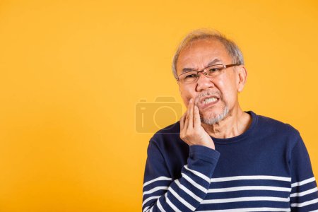 Téléchargez les photos : Douleur dentaire. Asiatique malheureux homme âgé problèmes avec gencive douleur studio tourné isolé sur fond jaune, Portrait vieil homme âgé triste main toucher la joue souffrant de maux de dents, soins dentaires - en image libre de droit
