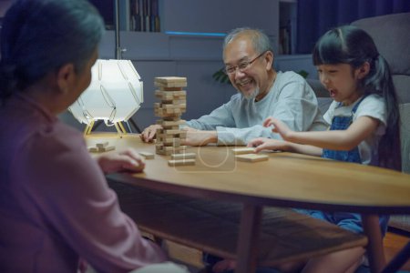 Photo for Happy grandparents Asian family enjoy playing toy block with little daughter and mother together at home night time, Smiling parent having fun play build constructor tower of wooden blocks, education - Royalty Free Image