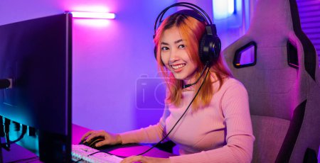 Téléchargez les photos : Happy Gamer endeavor plays online video games tournament with computer neon light, Smiling young woman wearing gaming headphones intend to do playing live stream games online at home looking to camera - en image libre de droit