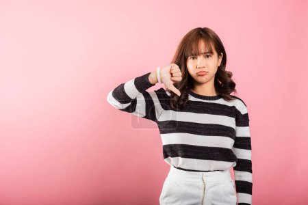 Foto de Una mujer asiática en un retrato mostrando su insatisfacción con un gesto de pulgares hacia abajo, señalando aversión. Estudio grabado aislado sobre un fondo rosa, capturando su expresión negativa. - Imagen libre de derechos