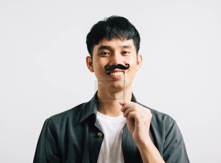 Téléchargez les photos : Un Asiatique joyeux dans un portrait tient une drôle de carte de moustache, rayonnant de bonheur et d'humour. Isolé sur le blanc, c'est une fête des Pères et une image sur le thème de novembre. - en image libre de droit