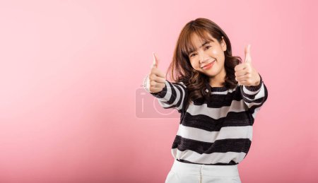 Téléchargez les photos : Une Asiatique souriante dans un portrait en studio sur fond rose fait un signe Ok pouce vers le haut, symbolisant l'accord et le succès. Son expression joyeuse rayonne de positivité et de satisfaction. - en image libre de droit