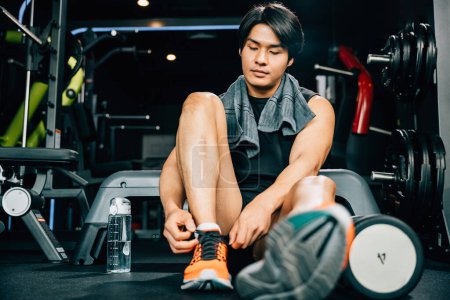 Photo for A muscular young man is tying his shoelaces on the gym floor, surrounded by dumbbells and other equipment. The shot emphasizes his strength and endurance during exercise - Royalty Free Image