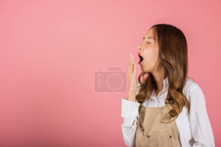 Foto de Retrato Asiática hermosa joven mujer usando overoles emociones cansado y soñoliento su bostezo cubriendo la boca abierta a mano, estudio de tiro aislado sobre fondo rosa, femenino atractivo insomnio aburrido - Imagen libre de derechos