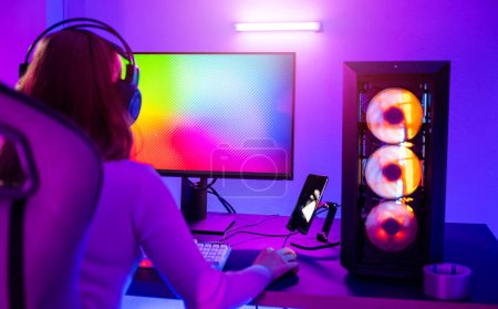 Photo for Asian professional gamer playing online video game on desktop computer PC have colorful neon LED lights, young woman in gaming headphones using computer for playing game at home, back view - Royalty Free Image