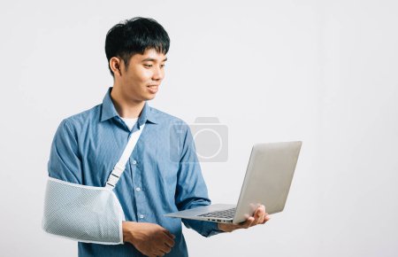 Téléchargez les photos : Homme d'affaires asiatique confiant, avec un bras cassé dans un bandage, travaille sur un ordinateur portable. Studio tourné isolé sur blanc, montrant sa détermination et son parcours de récupération. - en image libre de droit