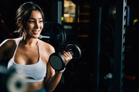 Photo for A smiling Asian woman in sportswear, lifting weights and holding a dumbbell, showcases her athletic biceps and strong body for a fitness portrait in the gym - Royalty Free Image
