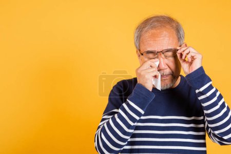 Téléchargez les photos : Aîné asiatique pleurer lever des lunettes avec tissu essuyer yeux rouges studio tourné isolé sur fond jaune, Portrait vieil homme âgé triste essuyer ses larmes, bouleversé déprimé solitaire - en image libre de droit