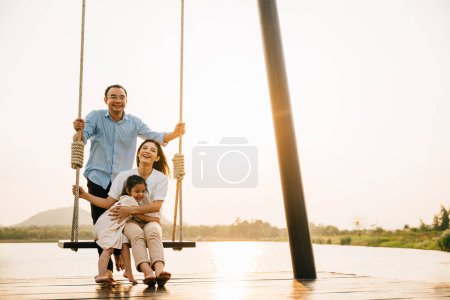 Foto de Una familia feliz disfrutando de un día soleado en el parque infantil, con el padre sentado y empujando el columpio mientras la madre y su hija juegan juntas, Feliz Día de la Familia - Imagen libre de derechos