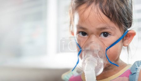Photo for Kid girl making makes inhalation nebulizer steam sick cough at home, Asian Child using nebulizer mask equipment alone have smoke, stuffy nose and runny, oxygen spray inhaler therapy, Health medical - Royalty Free Image