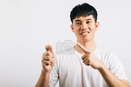 Foto de Retrato de un adolescente asiático sonriente sosteniendo un cepillo de dientes, cepillándose los dientes y señalándolo con alegría. Concepto de salud dental en un estudio filmado aislado en blanco, promoviendo el cuidado bucal. - Imagen libre de derechos