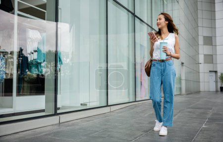 Foto de Estilo de vida moderno: Mujer sosteniendo una taza de vaso y navegando por su teléfono inteligente mientras camina por una bulliciosa calle de la ciudad - Imagen libre de derechos