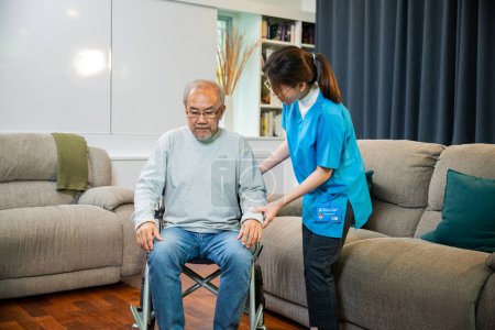 Foto de Asian nurse assisting helping senior man patient get up from wheelchair for practice walking at home, Smiling doctor support old man to getting up, help handicapped elderly stand up, healthcare - Imagen libre de derechos
