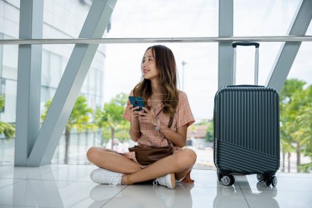 Téléchargez les photos : Femme asiatique assis sur le sol avec ses bagages et en utilisant son téléphone pour rester connecté tout en voyageant. Voyage touristique concept de voyage - en image libre de droit