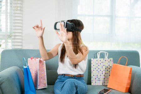 Photo for Happy young woman customer wearing 3d VR glasses virtual reality headset with shopping bags around she touching and pointing on air to online shopping in living room at home, digital cyberspace - Royalty Free Image