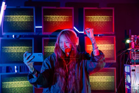 Photo for Winning. Confused Asian woman wearing gaming headphones feeling excited at gaming room, Happy Gamer playing video game online with smart mobile phone with neon lights raises hands to wins celebrating - Royalty Free Image