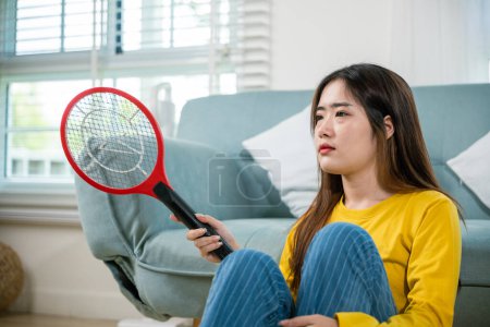 Téléchargez les photos : Asiatique femme assise à l'aide d'un dispositif de moustique swatter ou raquette de filet électrique, Jeune femme tuant les moustiques main tenant batteuse comme une arme dans le salon à la maison, tueur d'insectes de la technologie - en image libre de droit