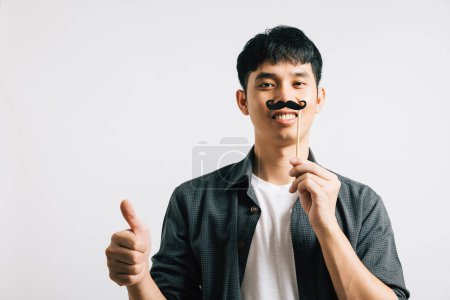 Téléchargez les photos : Un portrait d'homme asiatique présente une expression ludique alors qu'il tient une drôle de carte de moustache, rayonnant de bonheur et d'humour. Isolé sur blanc pour la Fête des Pères et le concept de novembre. - en image libre de droit