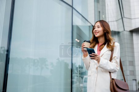 Téléchargez les photos : Femme d'affaires efficace maîtrisant le multitâche, tenant café et smartphone, restant productive sur son chemin vers le travail. - en image libre de droit
