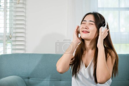 Foto de Mujer joven asiática que usa auriculares escuchando música con un teléfono móvil inteligente y relajándose mientras está sentada en el sofá en la sala de estar en casa, mujer que usa auriculares escucha música en el teléfono inteligente - Imagen libre de derechos
