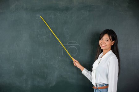 Foto de Back to school concept. Asian female teacher smiling with wooden stick pointing to blackboard at school in classroom, Happy beautiful young woman standing hold pointer to back board - Imagen libre de derechos