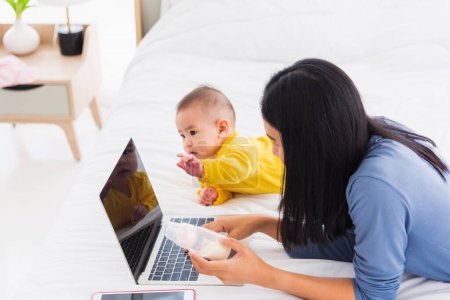 Téléchargez les photos : Asiatique nouvelle jeune belle mère trouver et lit des informations sur un ordinateur portable pour le mélange de lait sur une bouteille dans la chambre à coucher à la maison - en image libre de droit