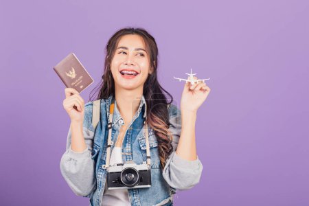Téléchargez les photos : Heureux portrait asiatique belle jeune femme excitée souriant en vacances d'été tenant jouet avion, passeport et appareil photo vintage, Thai voyage prêt féminin isolé sur fond violet - en image libre de droit