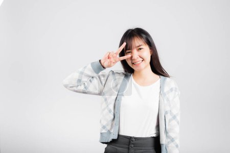 Téléchargez les photos : Woman showing finger making v-sign victory symbol near eye looking to camera, Happy Asian beautiful young female mark peace gesture symbol, studio shot isolated on white background with copy space - en image libre de droit
