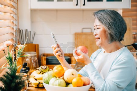 Photo for In the kitchen, an old Asian woman, a grandmother, enjoys her breakfast. She smiles while using her smartphone and eating a red apple. A portrayal of modern technology and happiness. - Royalty Free Image
