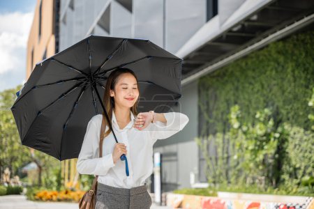 Foto de Una joven mujer de negocios asiática, protegida por un paraguas, camina a la oficina bajo la luz del sol, comprobando la hora durante un ajetreado día de trabajo. Su compromiso con el éxito es evidente en su mirada decidida. - Imagen libre de derechos