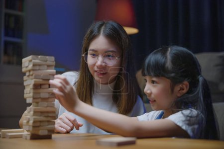 Photo for Asian young mother playing game in wood block with little daughter in home living room at night, Smiling woman help teach preschooler kid play build constructor tower of wooden blocks, family funny - Royalty Free Image