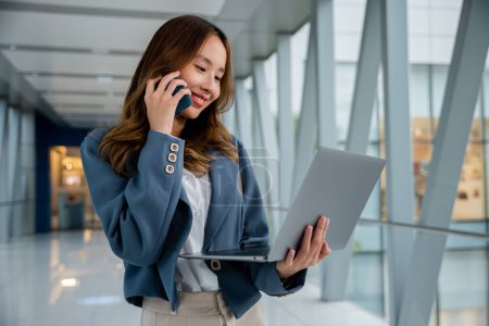 Téléchargez les photos : Une jeune femme d'affaires indépendante asiatique confiante qui regarde un ordinateur portable avec le sourire tout en utilisant un smartphone et en travaillant à l'extérieur dans la ville. Concept de travail à distance dans le bureau. - en image libre de droit