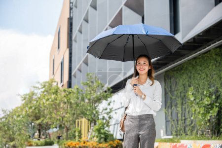 Foto de En el clima sofocante, una joven empresaria sostiene un paraguas mientras camina hacia la oficina. Su expresión severa y comportamiento exitoso son evidentes a pesar de la temperatura caliente. - Imagen libre de derechos