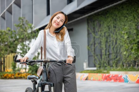 Foto de Una mujer de negocios con casco y alegre utiliza su teléfono inteligente mientras está de pie con su bicicleta al aire libre. Esta imagen moderna refleja el equilibrio exitoso de la alegría y la tecnología del trabajo. - Imagen libre de derechos