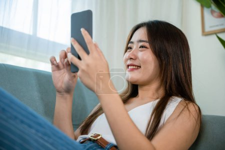 Téléchargez les photos : Happy Asian young woman checking social media holding smartphone at home while relaxing on sofa living room, Smile beautiful female sitting on couch using mobile phone to shopping online - en image libre de droit