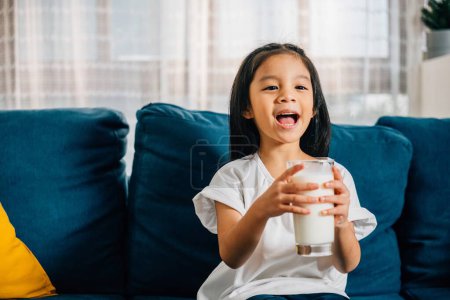 Téléchargez les photos : Un joli portrait d'un enfant asiatique tenant un verre de lait souriant de joie assis sur le canapé. Cette photo souligne l'importance de nourrir les enfants avec du lait riche en calcium. - en image libre de droit