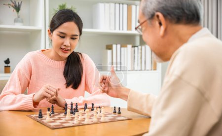Foto de Beautiful young smile woman having fun sitting playing chess game with senior elderly at home, nurse caregiver in nursing home for leisure, Happy active retired people, Healthcare and medical concept - Imagen libre de derechos