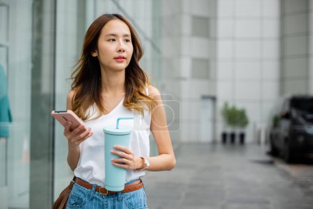 Photo for Young woman in white shirt holding smartphone and tumbler glass, enjoying a refreshing drink on a sunny city street. - Royalty Free Image