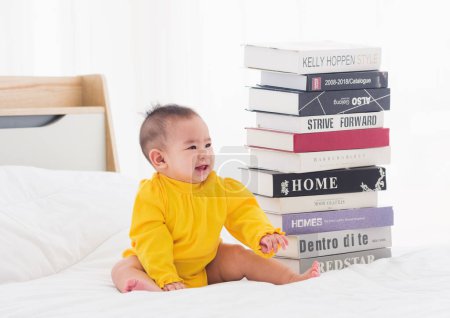Photo for Asian cute baby little girl toddler looking at a stack of books on bedroom white bed, First baby library concept - Royalty Free Image