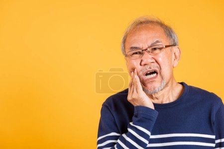 Téléchargez les photos : Dental pain. Portrait senior old man sad hand touching cheek suffering from toothache studio shot isolated on yellow background, Asian unhappy elder man problems with teeth pain, dental healthcare - en image libre de droit
