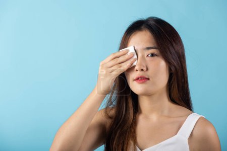 Téléchargez les photos : Facial Beauty Treatment Hygiene skin. Asian beautiful young woman removing cosmetic make up using pad cotton face cleaner disc, studio shot isolated on blue background, Happy female cleaning eyes - en image libre de droit