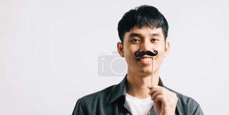 Téléchargez les photos : Un portrait d'homme asiatique capture son expression joyeuse tout en tenant une drôle de carte de moustache. Isolé sur blanc, il symbolise la joie et l'humour pour la fête des Pères et Novembre. - en image libre de droit