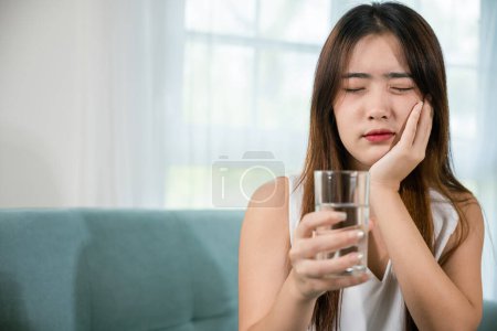 Téléchargez les photos : Asian young woman with sensitive teeth holding glass of cold water at home in living room, beautiful female toothache and dental problems touching cheek feeling pain after drinking cold water - en image libre de droit